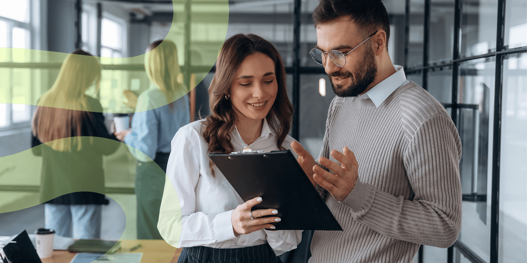 Two office workers are standing side by side, talking about something on a clipboard.